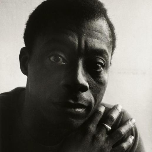 A black-and-white portrait of James Baldwin with his right hand on his left shoulder.