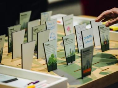 A hand hovers over a parking building interactive which includes small pieces of paper with words like park and lights in the to Live and Breathe exhibition.